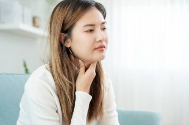 Asyalı kadın boğazı ağrıdığı için elini boynuna koydu. Dişi bademcik iltihabı iltihaba neden olur. Su alarak, antibiyotik alarak, doktora, virüse, korona virüsüne, gribe, gribe danışarak tedavi olunur.