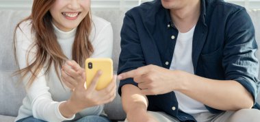 Gülümseyen Asyalı çift internet alışverişinde akıllı telefon kullanıyor. Koca, güzel bir eş için doğum günü hediyesi ödemeye hazır. Balayı için internetten konaklama, uygulama, uçak biletleri..