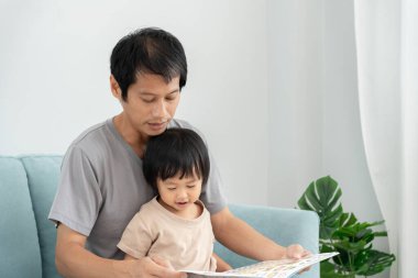 Mutlu Asyalı baba rahatla ve evde bebek zamanlarıyla kitap oku. Ebeveyn kızıyla kanepede oturup masal okuyor. öğrenme, çocuk bakımı, gülme, eğitim, hikaye anlatma, uygulama.