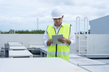 Asyalı bakım mühendisi fabrikanın çatısında çalışıyor. Müteahhit kompresör sistemini denetleyecek ve inşaatta havalandırma sistemi kurmayı planlıyor. teknoloji, çevrimiçi, mobil uygulama.