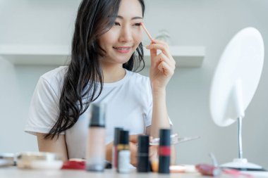 Güzel bir Asyalı kadın aynanın karşısına oturur ve makyaja gülümser. Makyaj yapan sağlıklı bir kadının yüzü. Reklam, yaşam tarzı, kozmetik, makyaj malzemeleri, güzellik aktiviteleri, güzellik