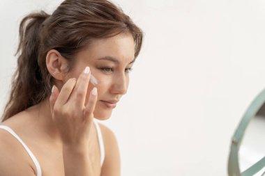 Güzel bir kadın aynanın önünde oturur. Krem sürerek makyaj yapan sağlıklı bir kadın yüzü. Cilt kremi reklamı, kırışıklık önleyici, bebek yüzü, beyazlatma, nemlendirici, gözenekleri sıkılaştırma serumu.