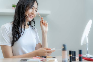 Güzel bir Asyalı kadın aynanın karşısına oturur ve makyaja gülümser. Makyaj yapan sağlıklı bir kadının yüzü. Reklam, yaşam tarzı, kozmetik, makyaj malzemeleri, güzellik etkinliği, güzellik uzmanı.