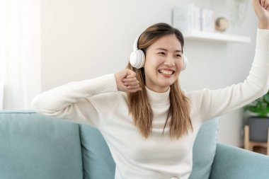 Kablosuz kulaklıklı neşeli genç Asyalı kadınlar oturup müzik dinliyor, eğleniyor, müzik dinliyor, kolunu kaldırıyor, en sevdiği şarkıyla dans ediyor, harika bir soundtrack 'in keyfini çıkarıyor, mutlu bir hayat sürüyor, eğleniyor.