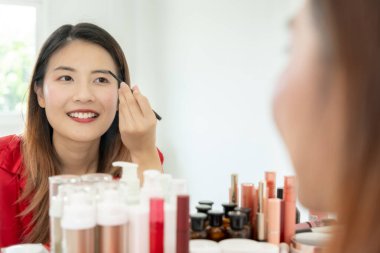 Güzel bir Asyalı kadın aynanın karşısına oturur ve makyaja gülümser. Makyaj yapan sağlıklı bir kadının yüzü. Reklam, yaşam tarzı, kozmetik, makyaj malzemeleri, güzellik etkinliği, güzellik uzmanı.