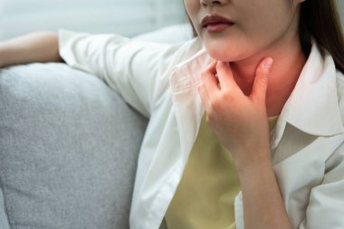 Asyalı kadın boğazı ağrıdığı için elini boynuna koydu. Dişi bademcik iltihabı iltihaba neden olur. Su alarak, antibiyotik alarak, doktora, virüse, korona virüsüne, gribe, gribe danışarak tedavi olunur.
