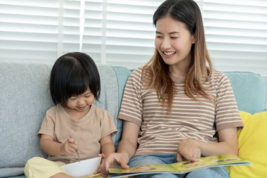 Mutlu Asyalı anne rahatla ve evde bebek zamanlarıyla kitap oku. Ebeveyn kızıyla kanepede oturup masal okuyor. öğrenme, çocuk bakımı, gülme, eğitim, hikaye anlatma, uygulama.