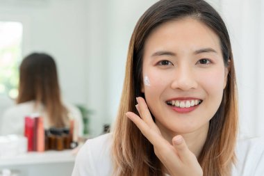 Güzel Asyalı kadın gülümsüyor, cilt kremi kullanıyor. Sağlıklı bir kadın yüzüne krem sürün ve makyaj yapın. Cilt kremi reklamı, kırışıklık önleyici, bebek yüzü, beyazlatma, nemlendirici, gözenekleri sıkılaştırma serumu.