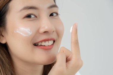 Güzel Asyalı kadın gülümsüyor, cilt kremi kullanıyor. Sağlıklı bir kadın yüzüne krem sürün ve makyaj yapın. Cilt kremi reklamı, kırışıklık önleyici, bebek yüzü, beyazlatma, nemlendirici, gözenekleri sıkılaştırma serumu.