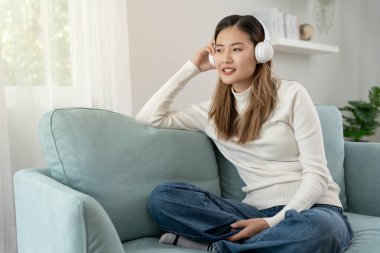 Kablosuz kulaklıklı neşeli genç Asyalı kadınlar oturup müzik dinliyor, eğleniyor, müzik dinliyor, kolunu kaldırıyor, en sevdiği şarkıyla dans ediyor, harika bir soundtrack 'in keyfini çıkarıyor, mutlu bir hayat sürüyor, eğleniyor.