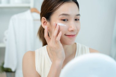 Güzel Asyalı kadın gülümsüyor, cilt kremi kullanıyor. Sağlıklı bir kadın yüzüne krem sürün ve makyaj yapın. Cilt kremi reklamı, kırışıklık önleyici, bebek yüzü, beyazlatma, nemlendirici, gözenekleri sıkma seru