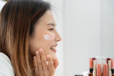 Güzel Asyalı kadın gülümsüyor, cilt kremi kullanıyor. Sağlıklı bir kadın yüzüne krem sürün ve makyaj yapın. Cilt kremi reklamı, kırışıklık önleyici, bebek yüzü, beyazlatma, nemlendirici, gözenekleri sıkılaştırma serumu.