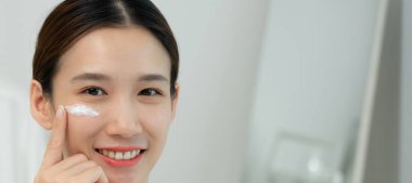 Güzel Asyalı kadın gülümsüyor, cilt kremi kullanıyor. Sağlıklı bir kadın yüzüne krem sürün ve makyaj yapın. Cilt kremi reklamı, kırışıklık önleyici, bebek yüzü, beyazlatma, nemlendirici, gözenekleri sıkılaştırma serumu.