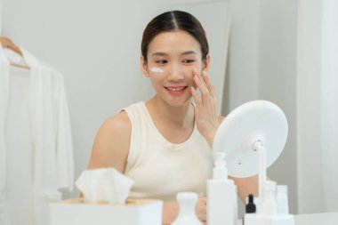 Güzel bir Asyalı kadın aynanın önünde oturur. Krem sürerek makyaj yapan sağlıklı bir kadın yüzü. Cilt kremi reklamı, kırışıklık önleyici, bebek yüzü, beyazlatma, nemlendirici, gözenekleri sıkılaştırma serumu.