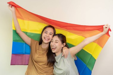 LGBT grubu. Güzel görünümlü lezbiyen çiftler gökkuşağı bayraklarını parlak bir şekilde örterler. Asyalı genç çift birbirlerine mutlu bir şekilde sarılıyorlar, aşık olan sevgili, biseksüeller, homoseksüellik, özgürlük, ifade, mutlu hayat.