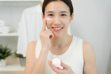 Güzel Asyalı kadın gülümsüyor, cilt kremi kullanıyor. Sağlıklı bir kadın yüzüne krem sürün ve makyaj yapın. Cilt kremi reklamı, kırışıklık önleyici, bebek yüzü, beyazlatma, nemlendirici, gözenekleri sıkılaştırma serumu.