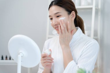 Güzel bir Asyalı kadın aynanın önünde oturur. Krem sürerek makyaj yapan sağlıklı bir kadın yüzü. Cilt kremi reklamı, kırışıklık önleyici, bebek yüzü, beyazlatma, nemlendirici, gözenekleri sıkılaştırma serumu.