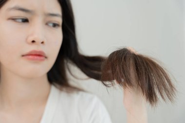 Asyalı kadın çok üzgün ve üzgün. Hasarlı saçlara, saç dökülmesine, saç dökülmesine, vitamin eksikliğine, kelliğe, doğum sonrası depresyona, çinko, regl veya endokrin bozukluklarına, hormonal dengesizliğe bakıyor.