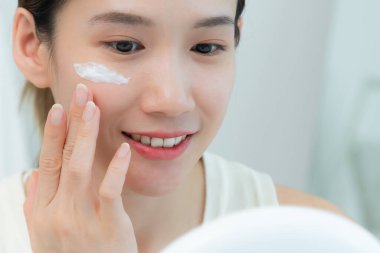 Güzel bir Asyalı kadın aynanın önünde oturur. Krem sürerek makyaj yapan sağlıklı bir kadın yüzü. Cilt kremi reklamı, kırışıklık önleyici, bebek yüzü, beyazlatma, nemlendirici, gözenekleri sıkılaştırma serumu.