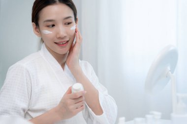 Güzel bir Asyalı kadın aynanın önünde oturur. Krem sürerek makyaj yapan sağlıklı bir kadın yüzü. Cilt kremi reklamı, kırışıklık önleyici, bebek yüzü, beyazlatma, nemlendirici, gözenekleri sıkılaştırma serumu.