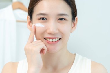 Güzel Asyalı kadın gülümsüyor, cilt kremi kullanıyor. Sağlıklı bir kadın yüzüne krem sürün ve makyaj yapın. Cilt kremi reklamı, kırışıklık önleyici, bebek yüzü, beyazlatma, nemlendirici, gözenekleri sıkılaştırma serumu.