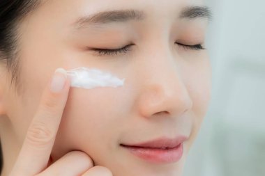 Güzel Asyalı kadın gülümsüyor, cilt kremi kullanıyor. Sağlıklı bir kadın yüzüne krem sürün ve makyaj yapın. Cilt kremi reklamı, kırışıklık önleyici, bebek yüzü, beyazlatma, nemlendirici, gözenekleri sıkılaştırma serumu.