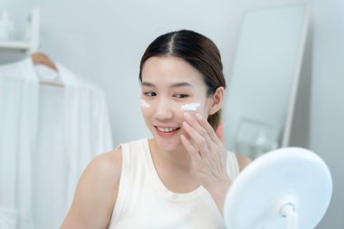 Güzel bir Asyalı kadın aynanın önünde oturur. Krem sürerek makyaj yapan sağlıklı bir kadın yüzü. Cilt kremi reklamı, kırışıklık önleyici, bebek yüzü, beyazlatma, nemlendirici, gözenekleri sıkılaştırma serumu.
