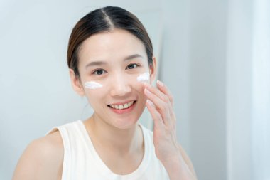 Güzel Asyalı kadın gülümsüyor, cilt kremi kullanıyor. Sağlıklı bir kadın yüzüne krem sürün ve makyaj yapın. Cilt kremi reklamı, kırışıklık önleyici, bebek yüzü, beyazlatma, nemlendirici, gözenekleri sıkma seru