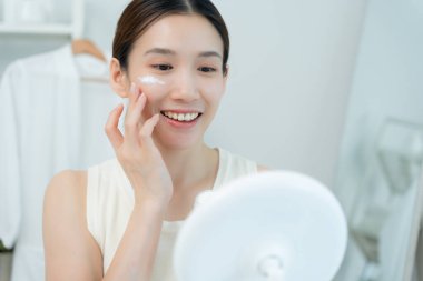 Güzel bir Asyalı kadın aynanın önünde oturur. Krem sürerek makyaj yapan sağlıklı bir kadın yüzü. Cilt kremi reklamı, kırışıklık önleyici, bebek yüzü, beyazlatma, nemlendirici, gözenekleri sıkılaştırma serumu.