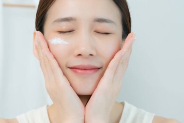 Güzel Asyalı kadın gülümsüyor, cilt kremi kullanıyor. Sağlıklı bir kadın yüzüne krem sürün ve makyaj yapın. Cilt kremi reklamı, kırışıklık önleyici, bebek yüzü, beyazlatma, nemlendirici, gözenekleri sıkılaştırma serumu.