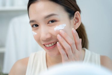 Güzel bir Asyalı kadın aynanın önünde oturur. Krem sürerek makyaj yapan sağlıklı bir kadın yüzü. Cilt kremi reklamı, kırışıklık önleyici, bebek yüzü, beyazlatma, nemlendirici, gözenekleri sıkılaştırma serumu.