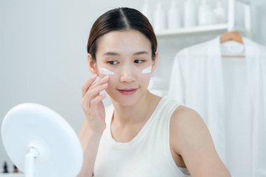 Güzel bir Asyalı kadın aynanın önünde oturur. Krem sürerek makyaj yapan sağlıklı bir kadın yüzü. Cilt kremi reklamı, kırışıklık önleyici, bebek yüzü, beyazlatma, nemlendirici, gözenekleri sıkılaştırma serumu.