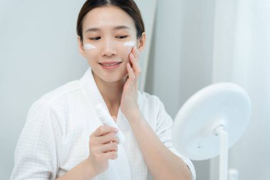 Güzel bir Asyalı kadın aynanın önünde oturur. Krem sürerek makyaj yapan sağlıklı bir kadın yüzü. Cilt kremi reklamı, kırışıklık önleyici, bebek yüzü, beyazlatma, nemlendirici, gözenekleri sıkılaştırma serumu.