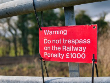 A sign with patchy, weathered red paint held by cable ties (one missing) to a metal gate warns the public not to trespass on the railway.