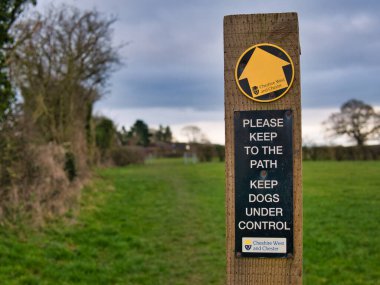 A way marker indicates a public footpath ahead, fixed to a wooden post in a field in winter in Cheshire, UK. A sign asks walkers to keep to the path and dogs on a lead.