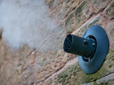 Water vapour from a gas central heating boiler flue condenses in cold air. The flue vents through the exterior wall of a sandstone building.