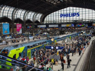 Hamburg, Germany - Jun 9 2024: Crowded, busy platforms at Hamburg Central Rail Station - Hauptbahnhof. A bright green RE7 regional express with double deck carriages stands at the platform. clipart