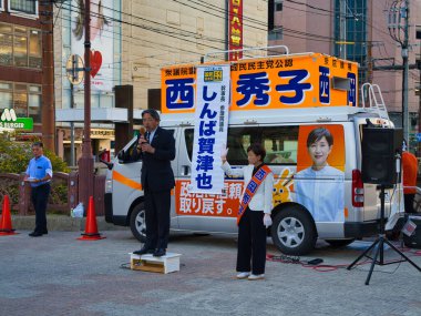 Nagasaki, Kyushu, Japonya 'da yerel politikacılar kampanya yapıyor. Bir konuşmacı, pankart ve hoparlörlerle süslenmiş bir minibüsün yanındaki platformdan kalabalığa sesleniyor..