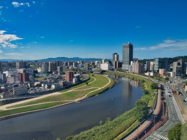 Kumamoto şehrinin ön planda Shirakawa Nehri ile hava manzarasında modern binalar, yeşil alanlar ve Japonya 'nın Kyushu adasında açık mavi gökyüzü sergileniyor..