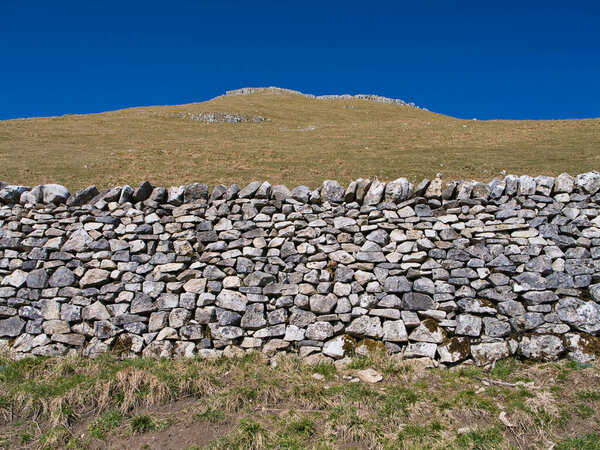 Dry stone wall in the Yorkshire Dales, constructed from irregularly shaped stones stacked without mortar, with a grassy hill rising gently towards the horizon under a clear blue sky.