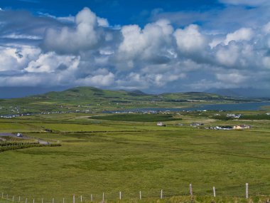 İrlanda 'nın Portmagee kentindeki dramatik bulutlu gökyüzü altında yuvarlanan yeşil tarlaların ve dağınık kır evlerinin panoramik görüntüsü.