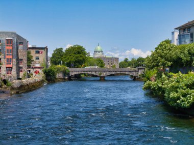 Galway, İrlanda - 13 Haziran 2025: İrlanda 'nın Galway kentindeki yemyeşil ve tarihi binalarla akan akan taştan bir köprünün görüntüsü.