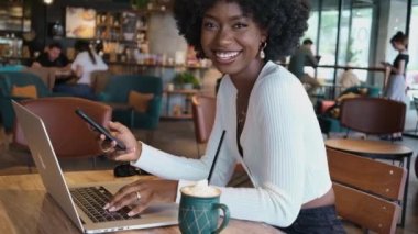 Kafede oturup kahve içen, dizüstü bilgisayarda ve telefonda çalışan mutlu Afrikalı Amerikalı kadın. Serbest çalışan iş kadını görüntülü mesaj yazıyor.