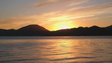 Renkli panoramik deniz manzarası ve turuncu renklerde bulutlu gün batımı gökyüzü, Akdeniz ve arka planda dağlar, suda günbatımı yansıması