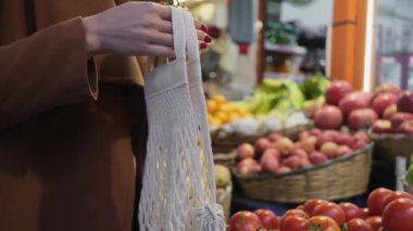 İp torbalı genç bir kadın marketteki sebze sebze rafından domates seçiyor sağlıklı besin maddeleri.