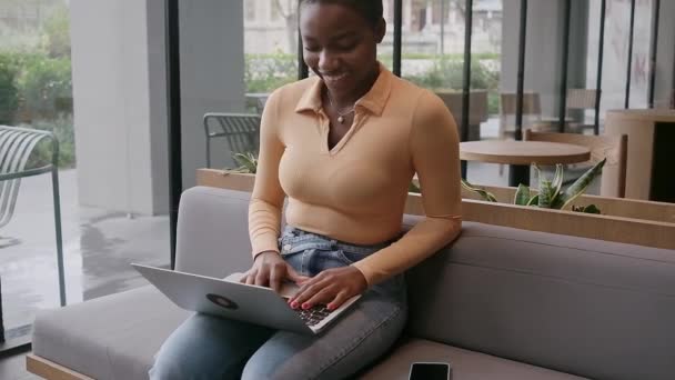 幸せなアフリカ系アメリカ人女性カジュアルな服装でカフェビジネスセンターに座っているか ラップトップや携帯電話で働いている大きな窓とのコーヒーを飲みながら フリーランスのビジネス女性のビデオ通話チャット入力メッセージ — ストック動画