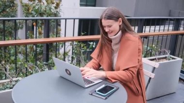 Güzel genç bir kadın iş merkezinin dışında oturuyor birlikte laptopta çalışıyor sonbahar gününde kazak ve palto giyerek daktilo yazıyor.