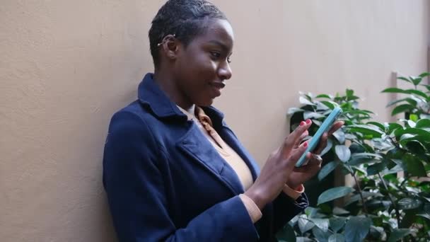 Афроамериканка Короткими Волосами Используя Телефон Снаружи Наклонись Стене Улыбаясь Прокрутка — стоковое видео