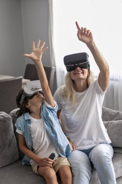 Anne ve oğlu sanal gerçeklik kulaklığı takıyor evdeki oturma odasında sanal gerçeklik oyunları oynayarak eğleniyorlar.