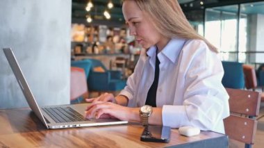 Beyaz gömlekli mutlu genç bir kadın kafeteryada oturuyor. Dizüstü bilgisayarda çalışıyor. Serbest çalışan bir iş kadını.